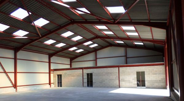 Entrepôt à ossature d'acier en construction ; charpente rouge, revêtement blanc, sol brun.