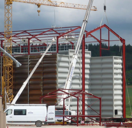 Intérieur d'un grand entrepôt avec poutres métalliques et toit translucide. La lumière du soleil pénètre à flots, illuminant l'espace vide.