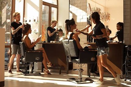 Zum Kontakt von Hairstylist by Yadenes. Zwei junge Frauen sitzen auf Stühlen vor Spiegeln im Friseursalon und bekommen von jeweils einem Friseur die Haare und Makeup gemacht.
