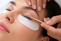Técnica aplicando extensiones de pestañas en el ojo cerrado de una mujer con almohadillas protectoras debajo de los ojos.