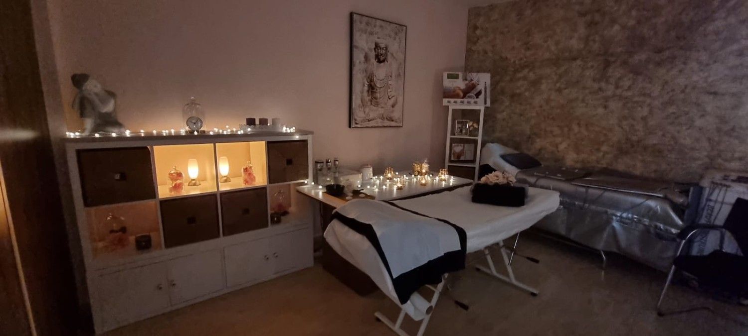Una sala de spa con iluminación tenue que cuenta con una camilla de masaje, un mueble de almacenamiento con luces de hadas y una pared de piedra texturizada como telón de fondo.