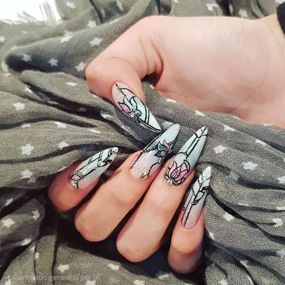 Uñas largas y puntiagudas en forma de almendra con intrincados diseños de líneas negras y delicados detalles florales en rosa, contrastando con una bufanda gris.