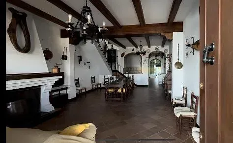 Amplio interior rústico: chimenea, escalera, mesa de comedor, lámparas de araña, vigas de madera, sillas.