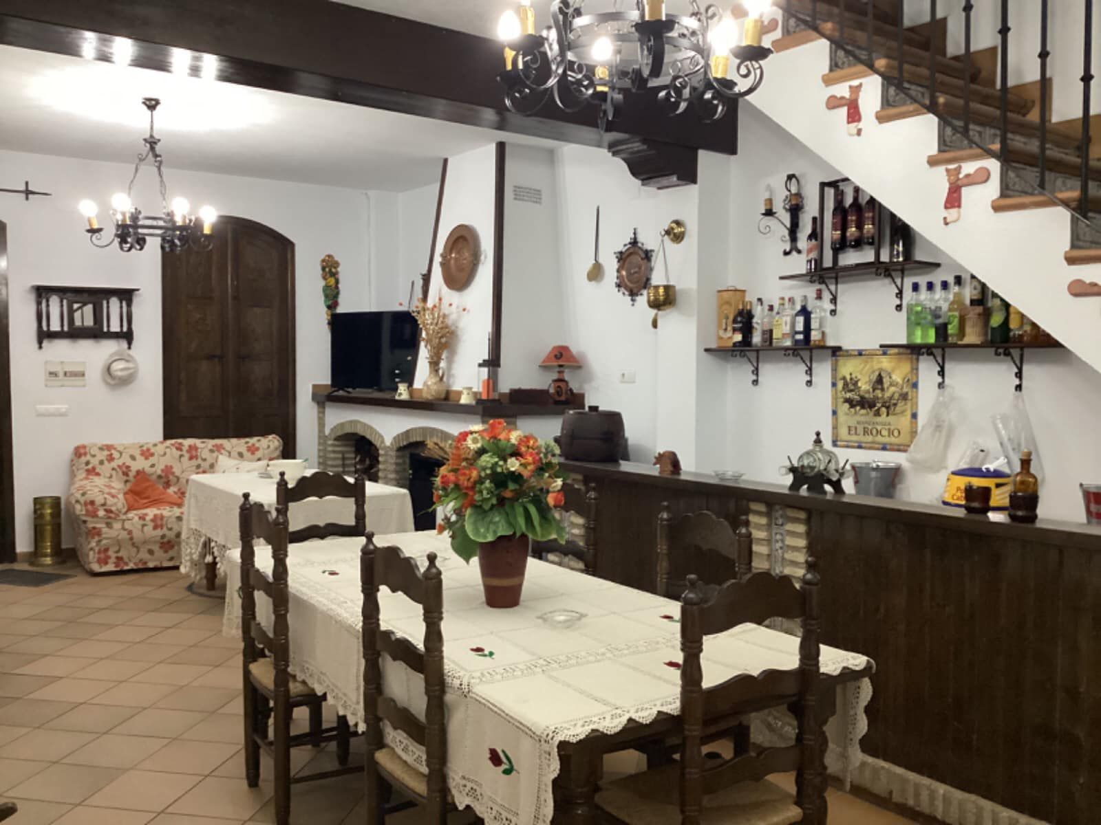 Comedor con mesa de madera, sillas y barra. Flores en la mesa, escalera y chimenea.