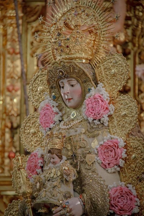 Una estatua de la Virgen María vestida de oro sosteniendo al Niño Jesús, adornada con rosas rosadas, dentro de una iglesia.
