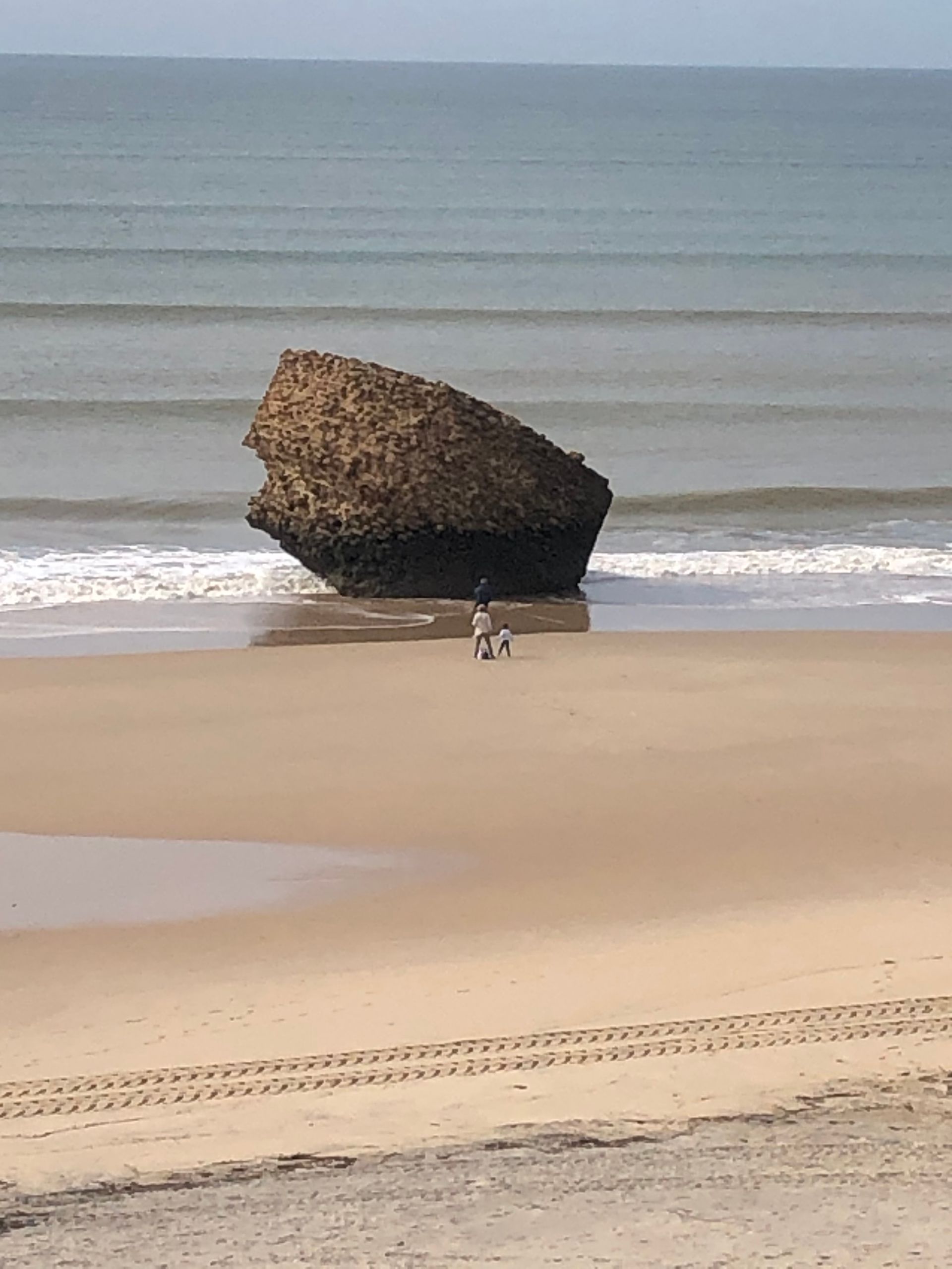 Escena de playa: gran roca en el agua, gente caminando en la arena, olas y un cielo azul.