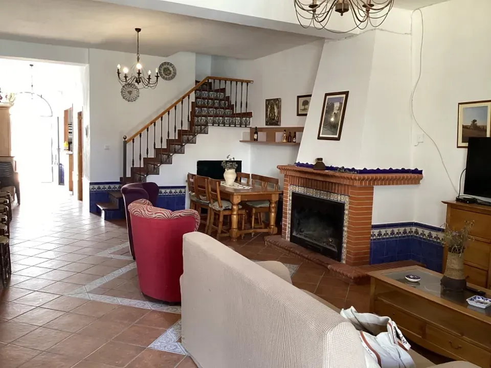 Amplia sala de estar con chimenea, escalera y comedor. Colores cálidos y suelo de baldosas.