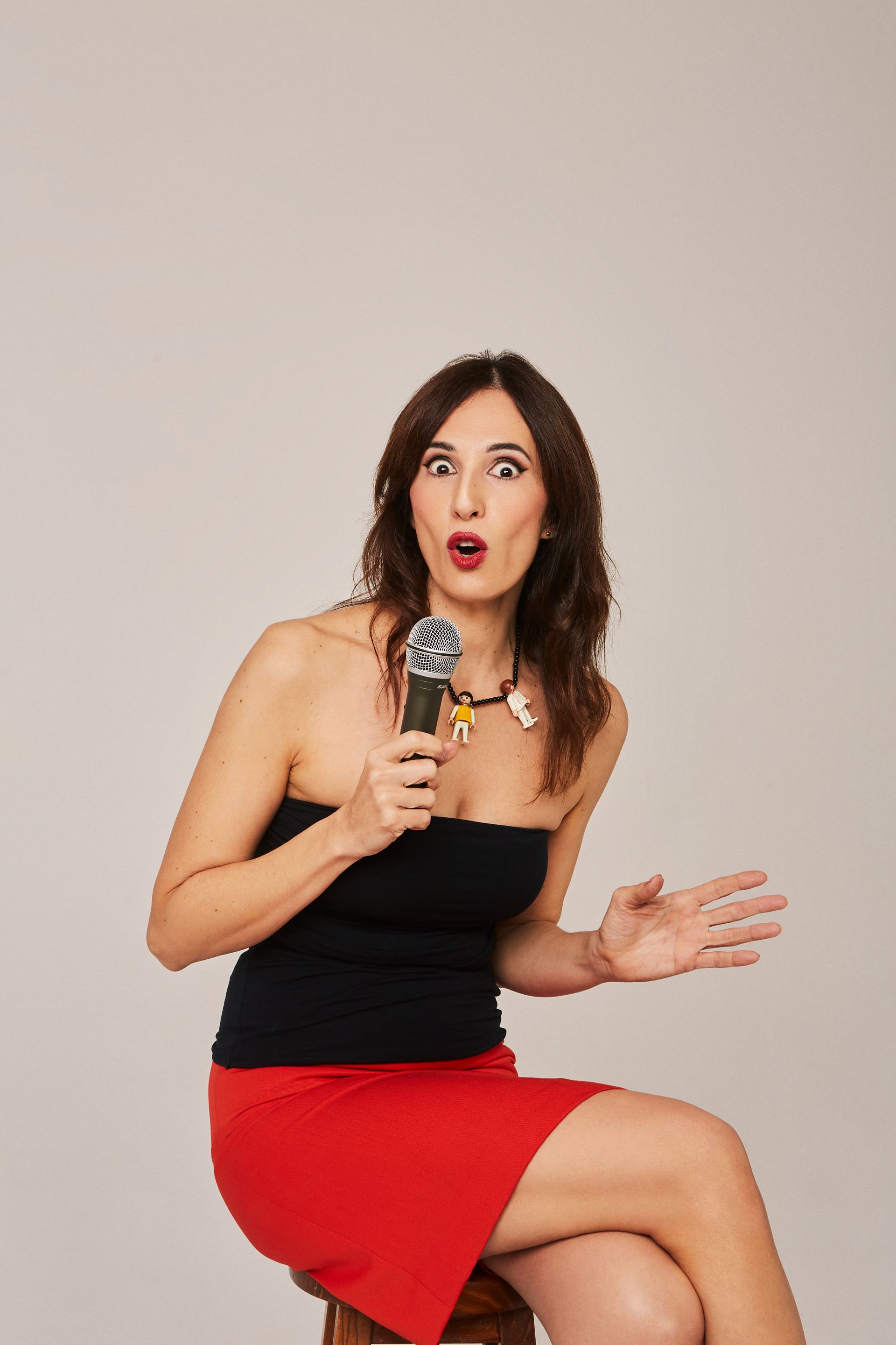 Mujer con expresión de sorpresa, sosteniendo un micrófono. Vestida con top negro y falda roja, sentada.