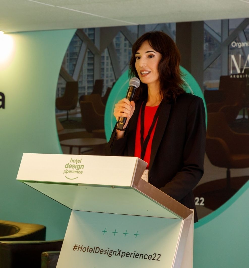 Mujer hablando en el podio en una conferencia; camisa roja, blazer negro, sosteniendo un micrófono, fondo de diseño de hotel.