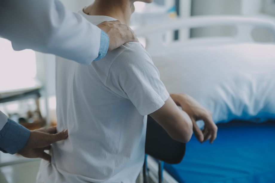 Médico examinando la espalda de un paciente en una habitación de hospital; batas blancas, camiseta blanca, cama al fondo.