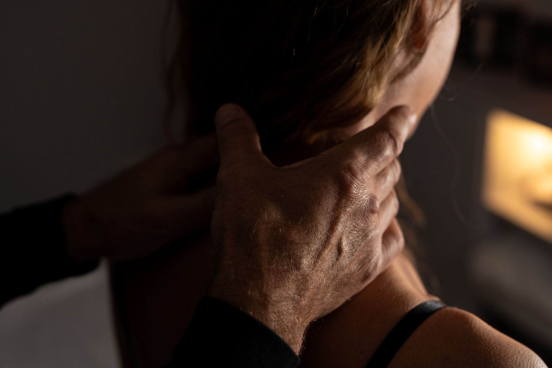 Manos que tocan suavemente el cuello de alguien por detrás, posiblemente durante un masaje. Poca luz, en interiores.
