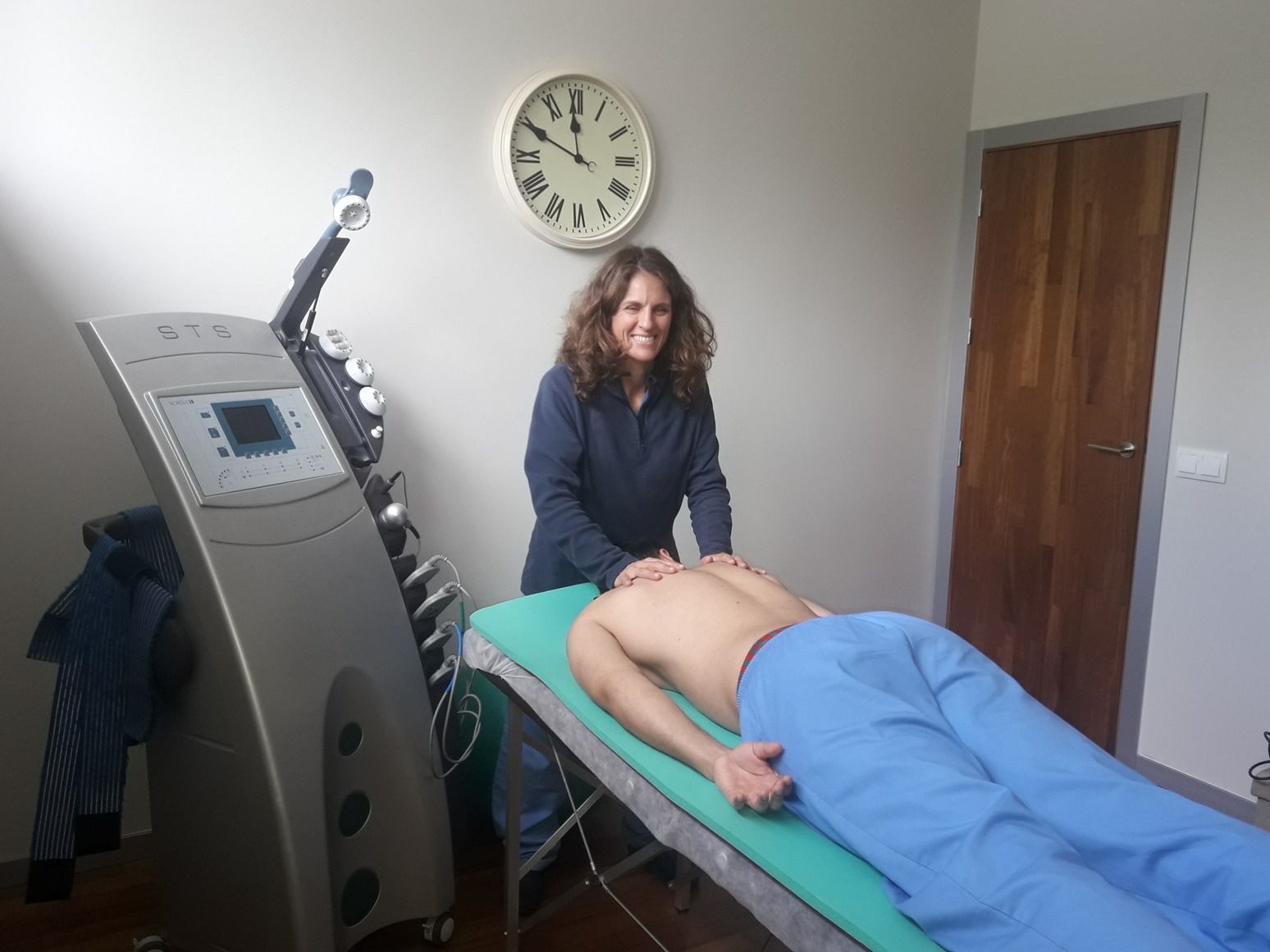 Mujer brindando tratamiento a un paciente acostado en una camilla; equipo médico al fondo.