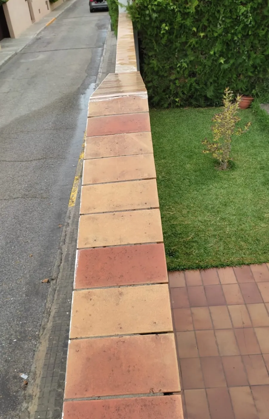 Acera junto a un jardín bordeado de ladrillos rojos, con césped a la derecha y una entrada para vehículos a la izquierda.