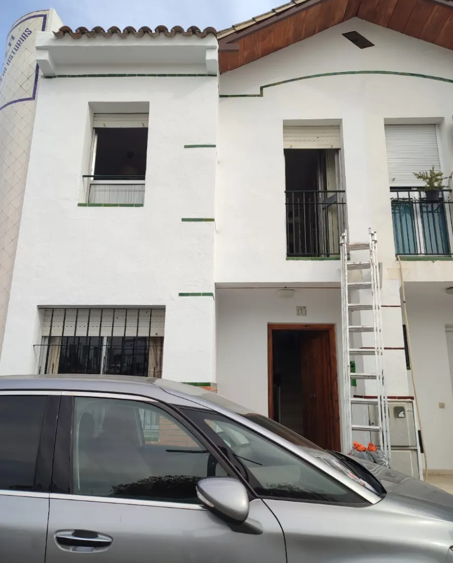 Casa blanca de dos plantas con una escalera en la fachada, parcialmente bloqueada por un coche plateado aparcado.