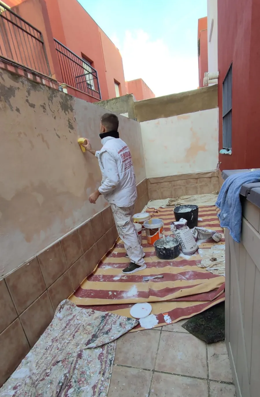 Persona pintando un pasillo exterior con paredes rosas; latas de pintura y herramientas sobre una alfombra estampada.