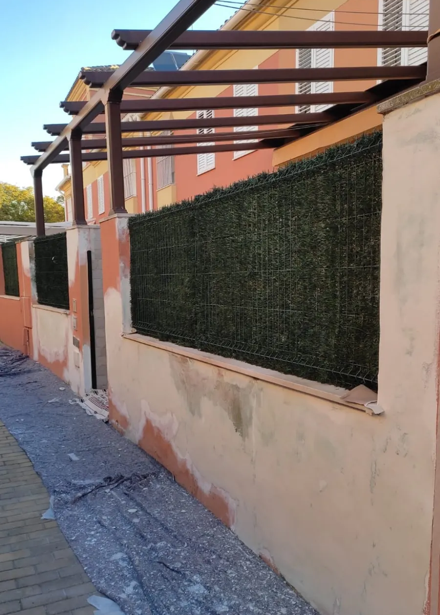 Acera estrecha junto a un muro color melocotón con un panel de seto verde oscuro y una pérgola metálica en la parte superior.