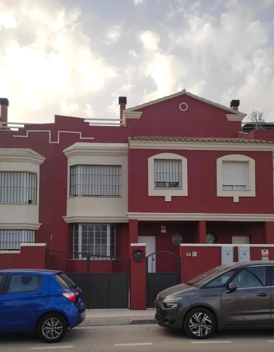 Casa adosada roja de dos plantas con detalles blancos y coches aparcados en una calle nublada.