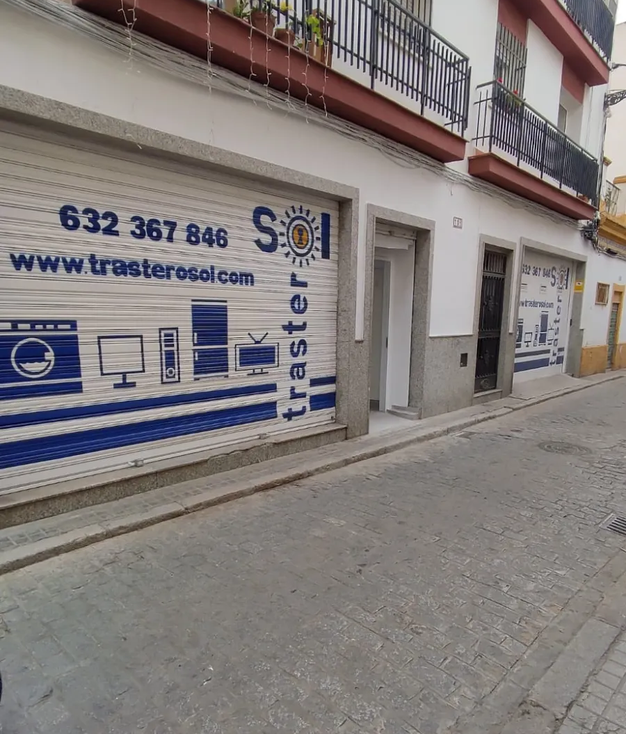 Vista de una callejuela empedrada estrecha con una tienda de fachada blanca y letreros azules bajo balcones rojos.
