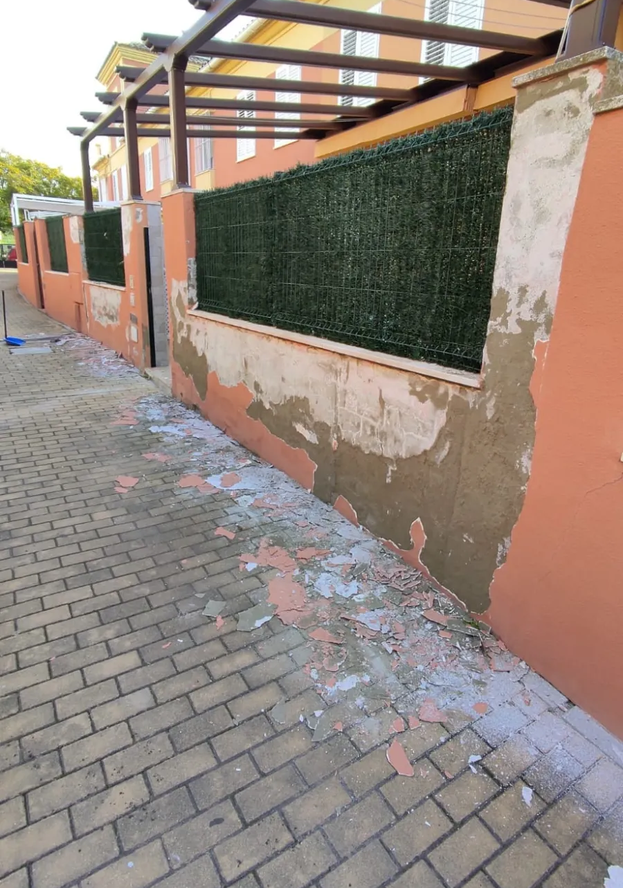 Muro de melocotón dañado y seto verde a lo largo de una acera de ladrillos bajo una valla de estructura metálica.