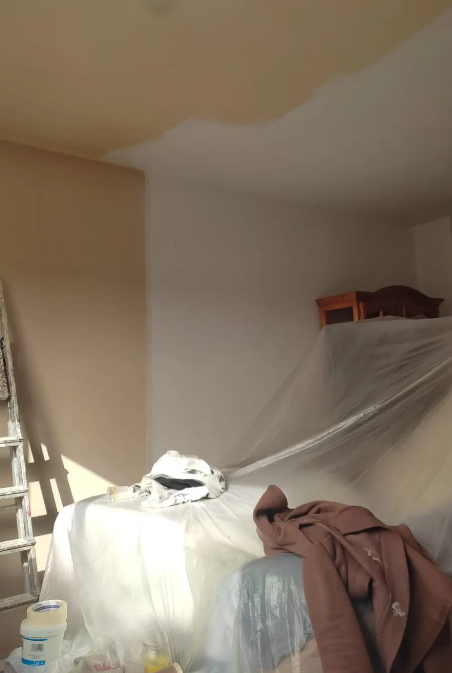 Habitación desordenada con ropa, una escalera y una cama cubierta con una lámina de plástico transparente bajo un techo dañado.
