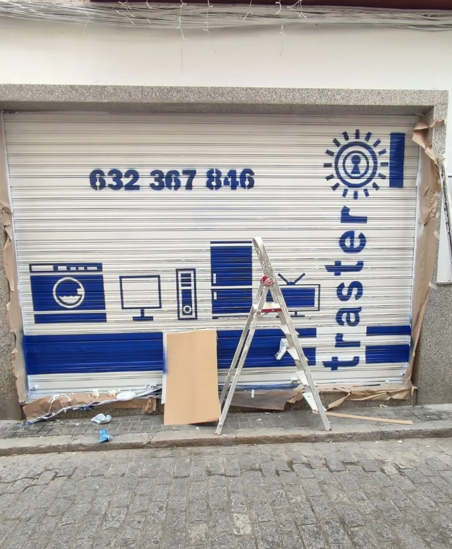 Fachada de una tienda con una persiana blanca, un letrero azul y una escalera apoyada contra ella, situada en la calle.