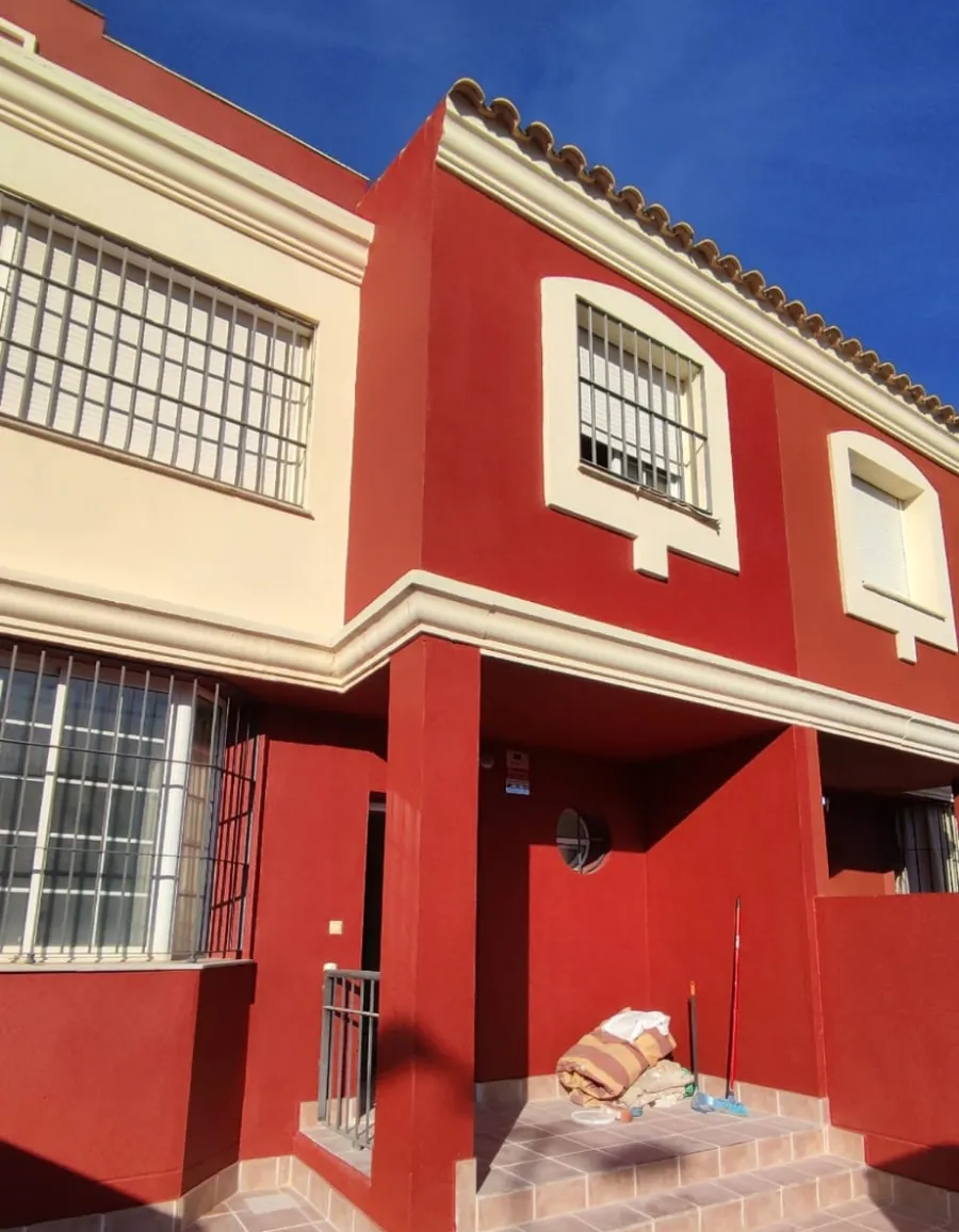 Casa adosada roja y crema con ventanas enrejadas bajo un cielo azul brillante, con un perrito en la entrada.