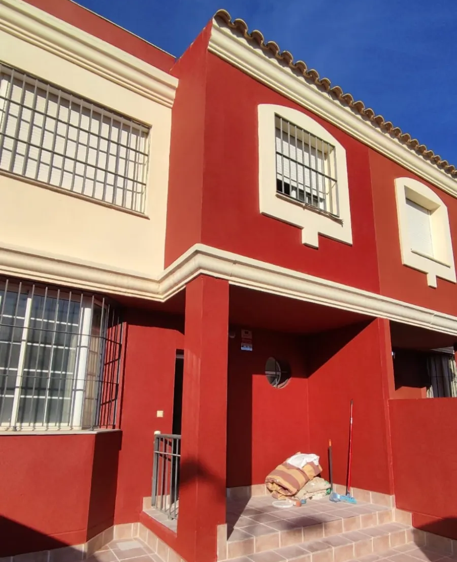 Exterior de una casa adosada roja y crema con ventanas enrejadas y una pequeña escalinata de entrada, bajo un cielo azul.