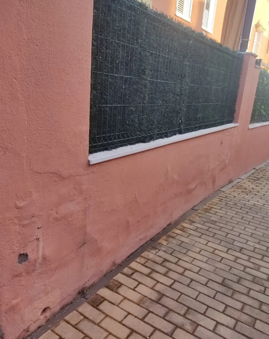 Muro rosa junto a una valla de malla negra y un sendero pavimentado con ladrillos en el exterior.
