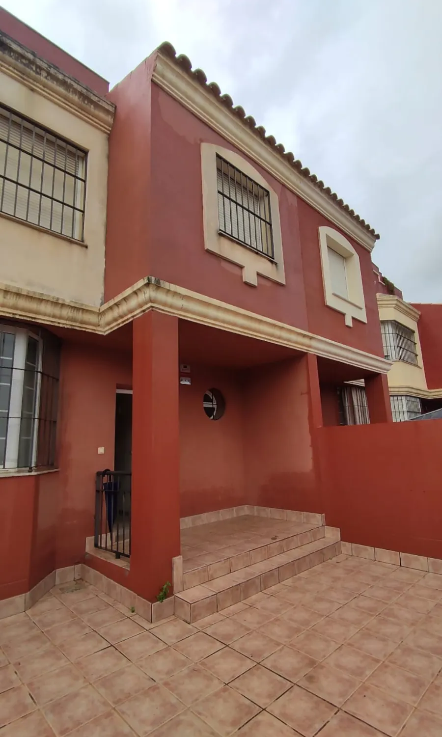 Casa roja de dos plantas con ventanas enrejadas, escalones de baldosas y un pequeño patio delantero.