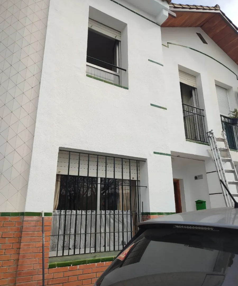Casa blanca de dos plantas con ventanas enrejadas y un coche negro aparcado delante.