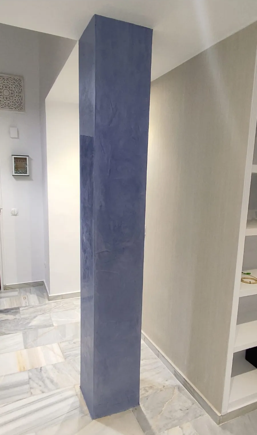 Columna cuadrada pintada de azul en un pasillo luminoso con suelo de baldosas y estanterías empotradas.
