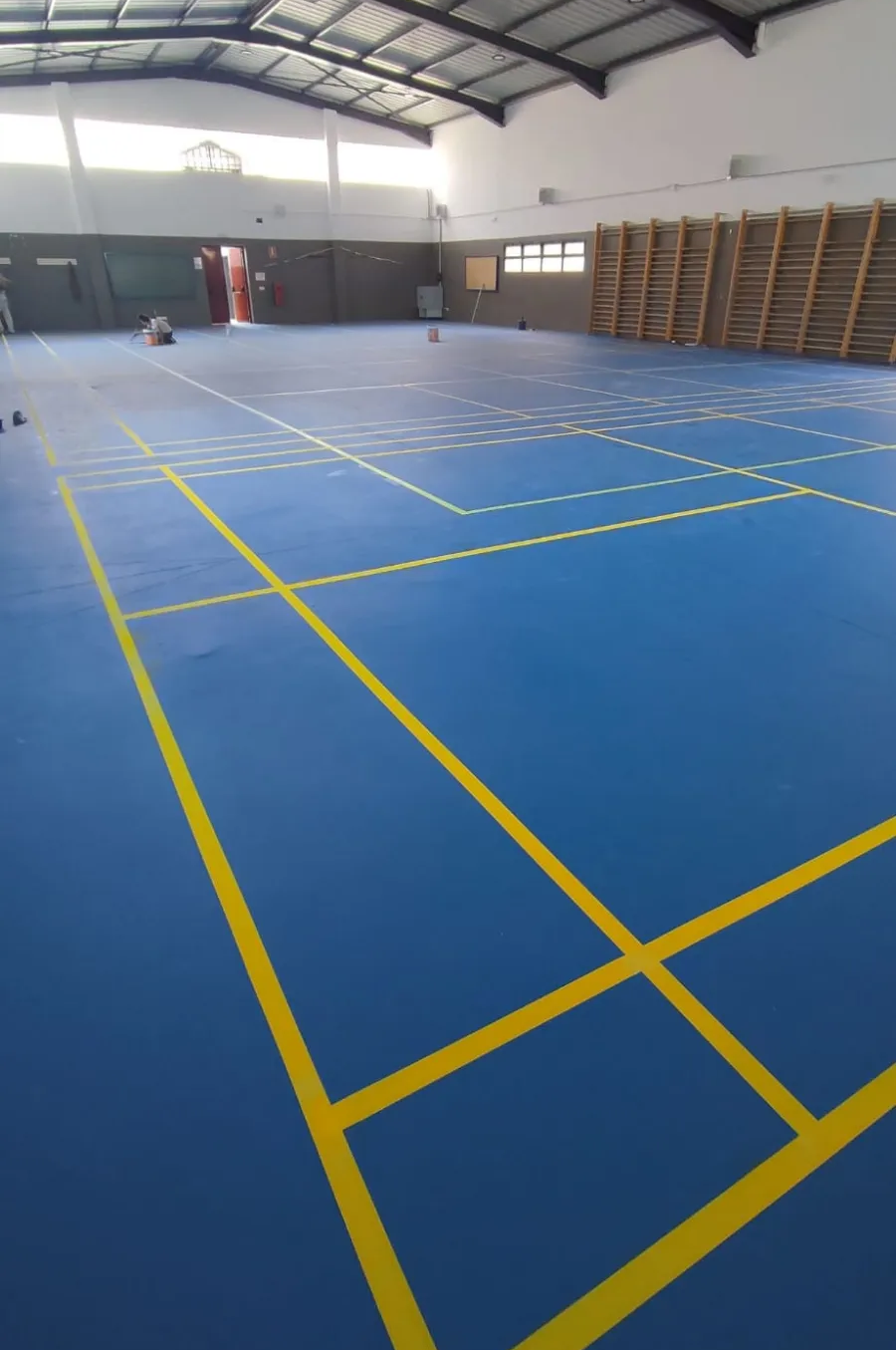 Pabellón deportivo cubierto con cancha azul, líneas amarillas y equipamiento apilado a lo largo de la pared derecha.