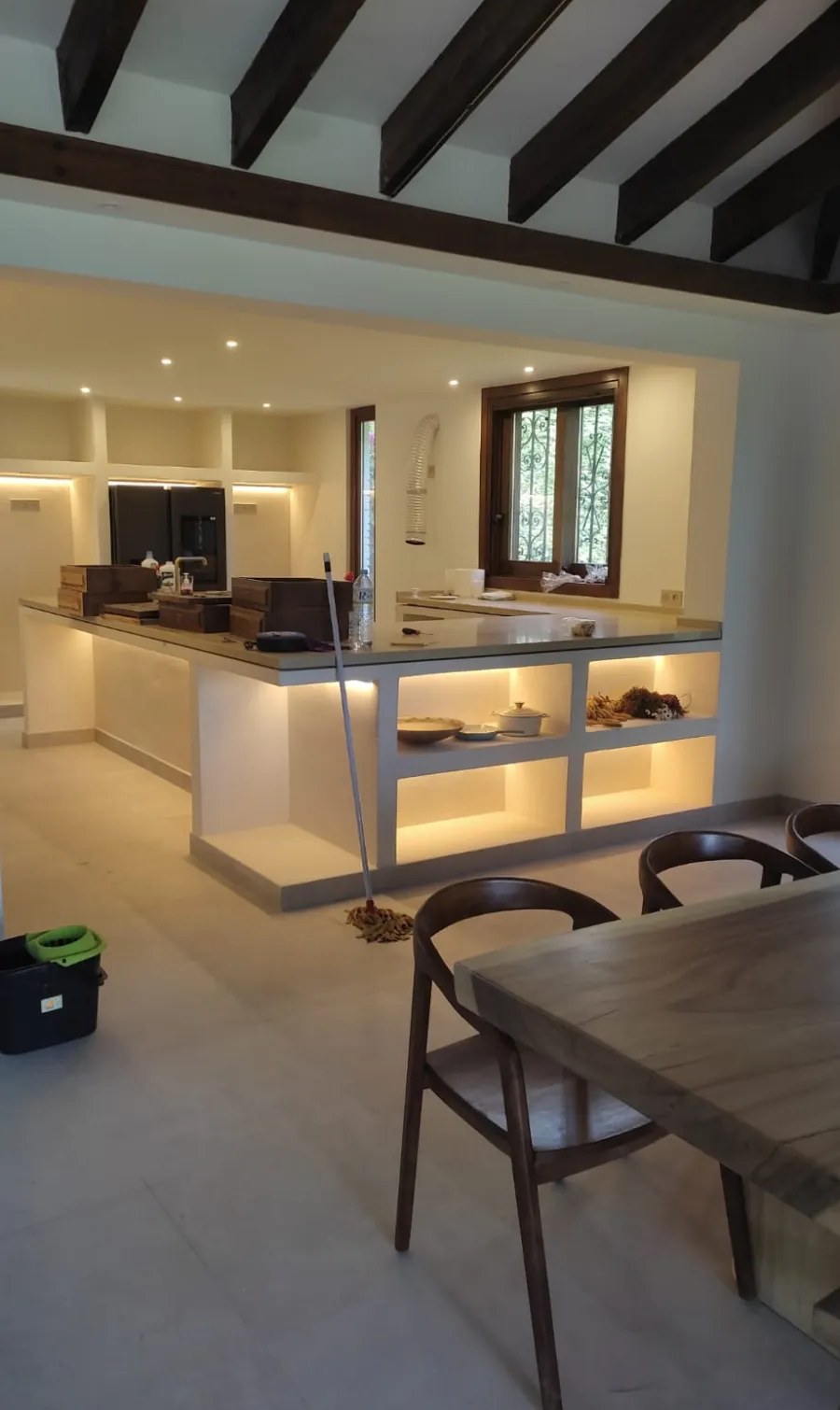 Espacio diáfano con vigas vistas, isla de cocina, mesa de comedor e iluminación empotrada cálida.
