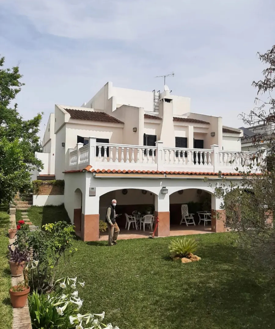 Casa blanca de dos plantas con balcón y porche, rodeada de césped y árboles.
