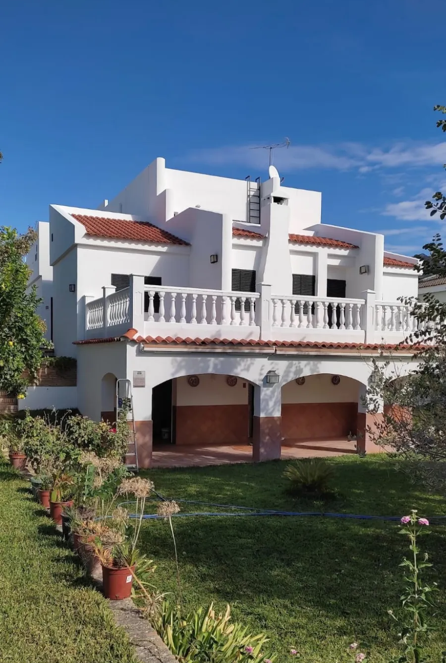 Casa blanca de dos plantas con detalles rojos en el tejado, balcones arqueados y un césped verde bajo un cielo azul.