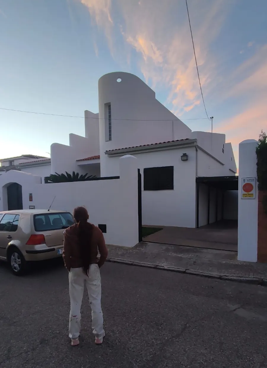 Una persona de pie en una calle frente a un edificio blanco al atardecer, con un coche aparcado cerca.
