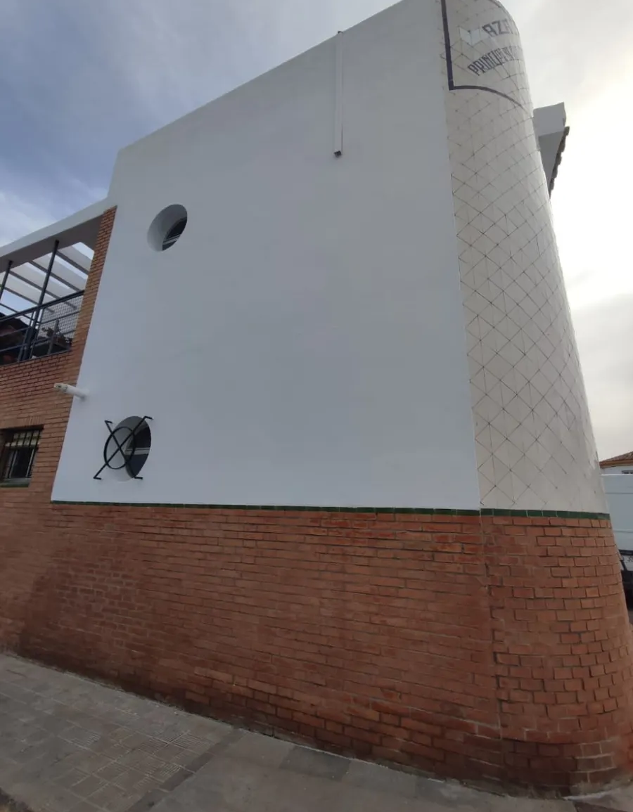 Edificio de estuco blanco con base de ladrillo y pequeñas ventanas redondas, visto desde abajo contra un cielo nublado.