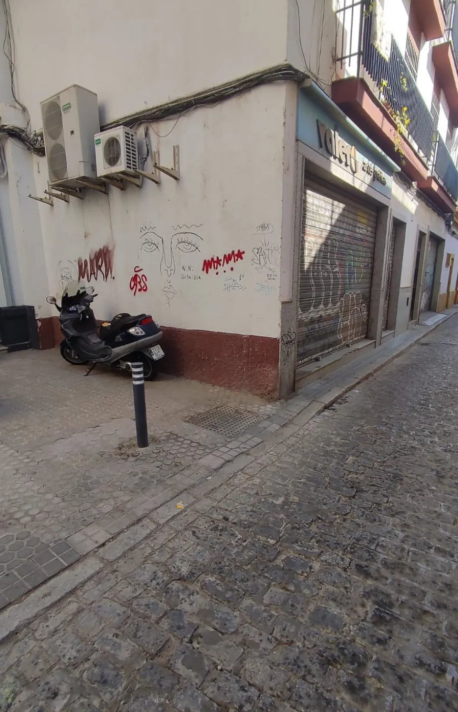 Esquina estrecha de una calle empedrada con un edificio blanco, una moto aparcada y locales comerciales cerrados.