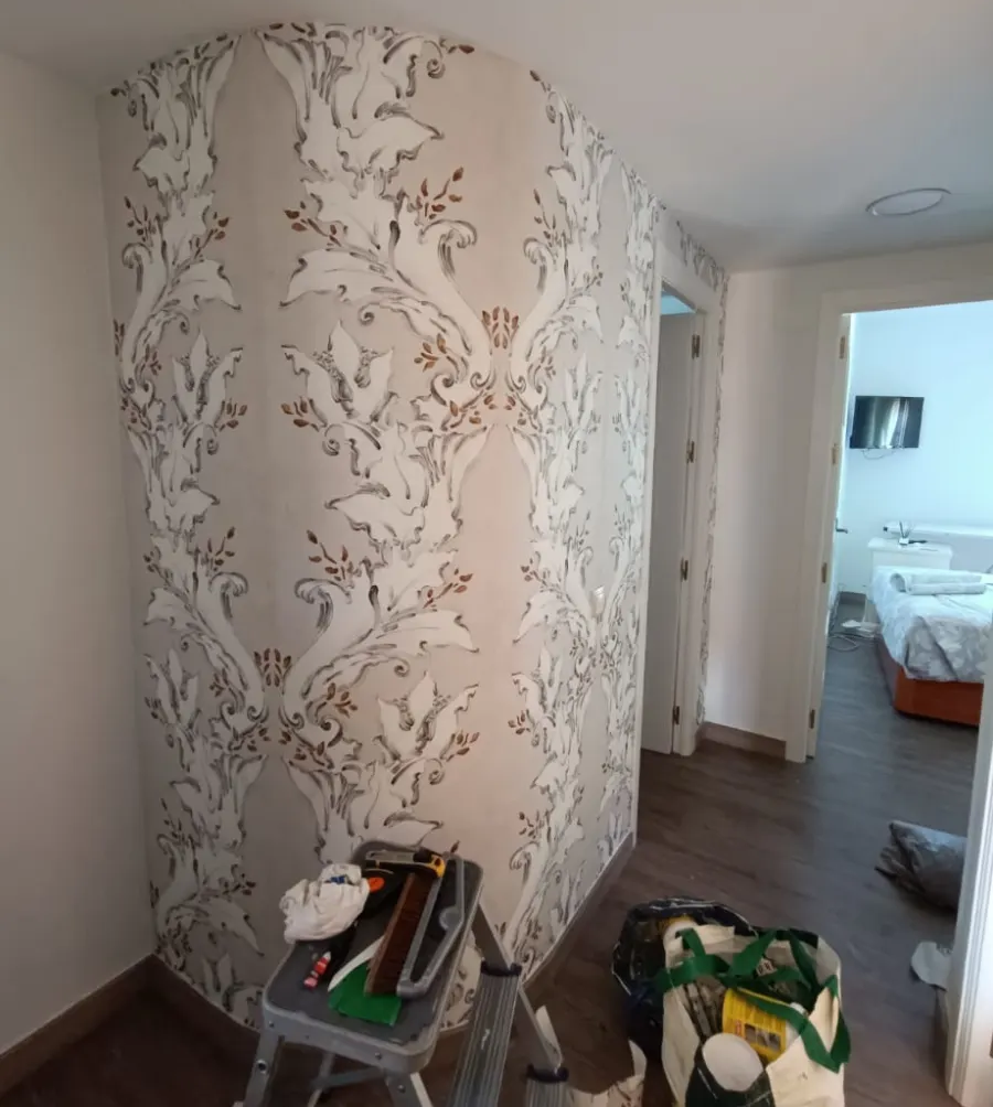 Pasillo con papel pintado floral, objetos apilados y una silla cerca de una puerta que da a un dormitorio.