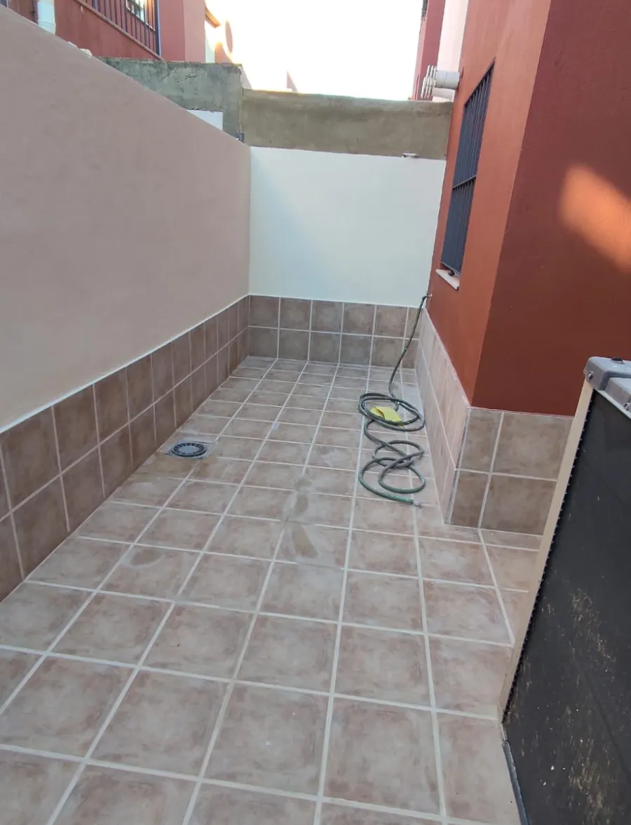 Patio estrecho con baldosas, un carrete para manguera, una pared blanca y paredes de edificio de color naranja.