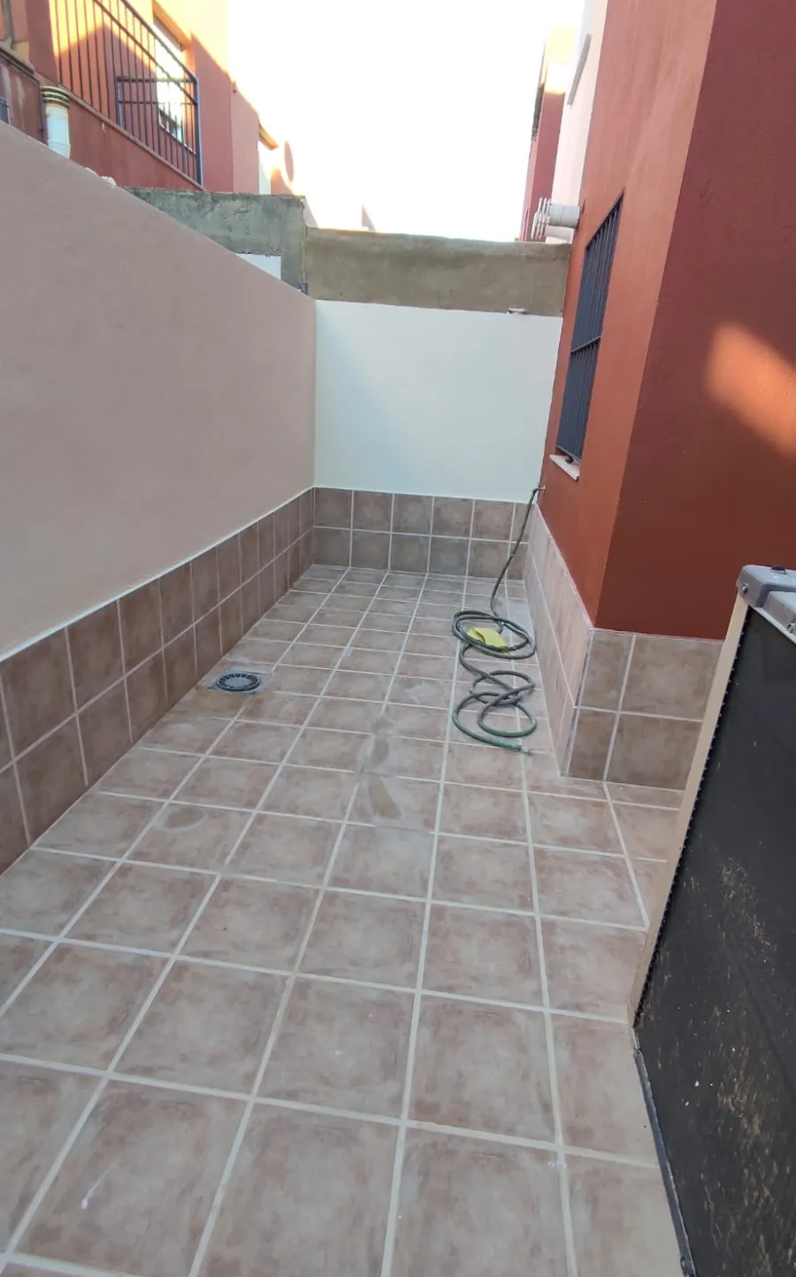 Estrecho pasillo exterior embaldosado con paredes blancas, laterales del edificio rojos y manguera enrollada en el suelo.