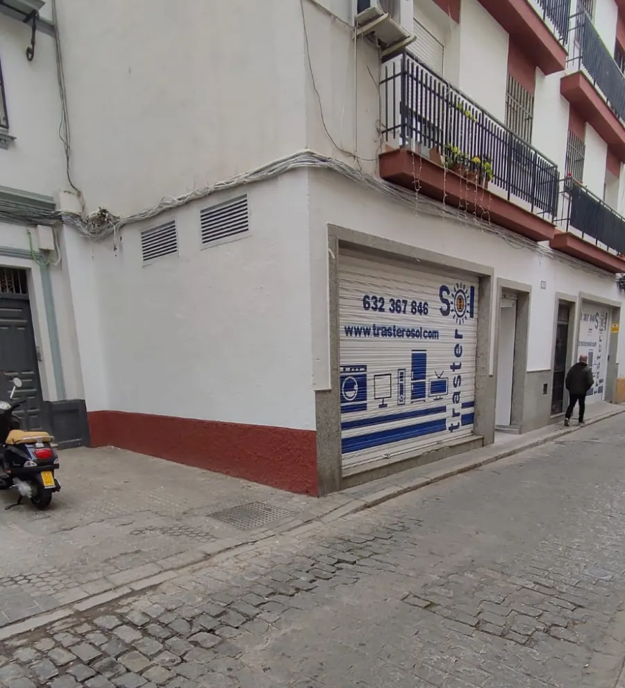 Esquina de una calle con un edificio blanco, detalles en rojo y un letrero en la acera, en una calle empedrada.