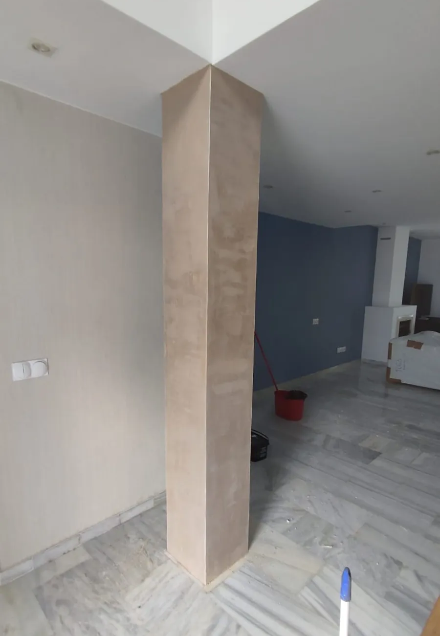 Habitación interior sin terminar con una columna cuadrada central, paredes de color beige y suelo de madera clara.