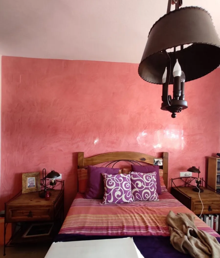 Habitación rosa con cama de madera, ropa de cama estampada, mesitas de noche y una gran lámpara colgante oscura.
