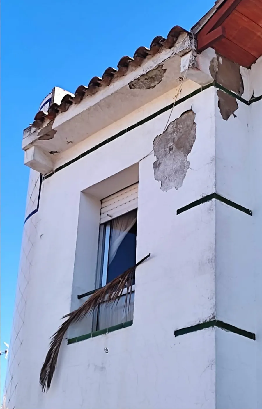 Fachada de edificio de estuco blanco con pintura descascarada, borde de tejado de tejas y ventana de balcón contra el cielo azul.