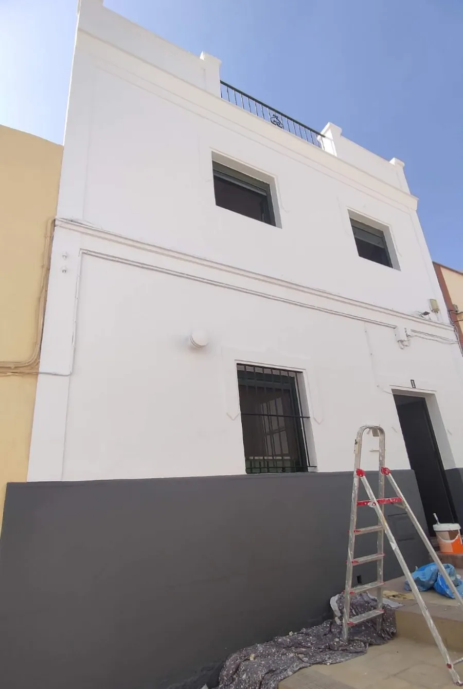 Exterior de una casa blanca de dos plantas con la pared inferior de color gris oscuro y una escalera apoyada contra la fachada.