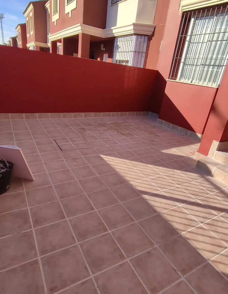Patio soleado con baldosas, paredes rojas y balcones de apartamentos, parcialmente sombreados por las sombras de las barandillas.