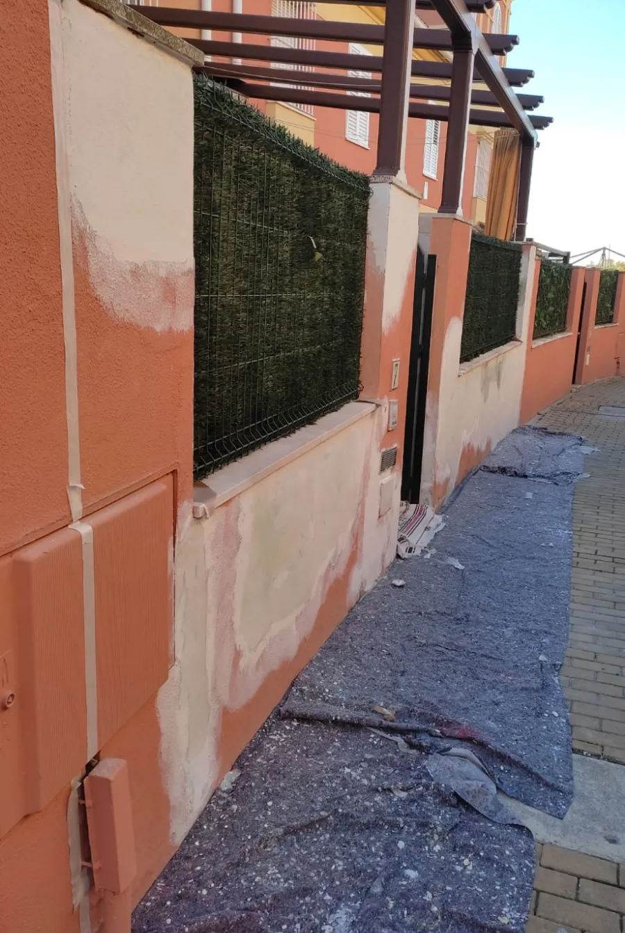 Pared naranja con paneles de malla verde a lo largo de un estrecho sendero pavimentado junto a un edificio.