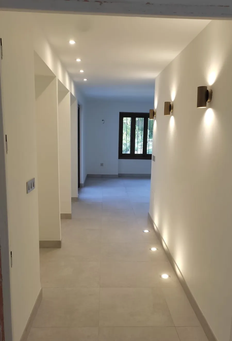 Pasillo luminoso con paredes blancas, apliques de pared, suelo de baldosas y una ventana al fondo.