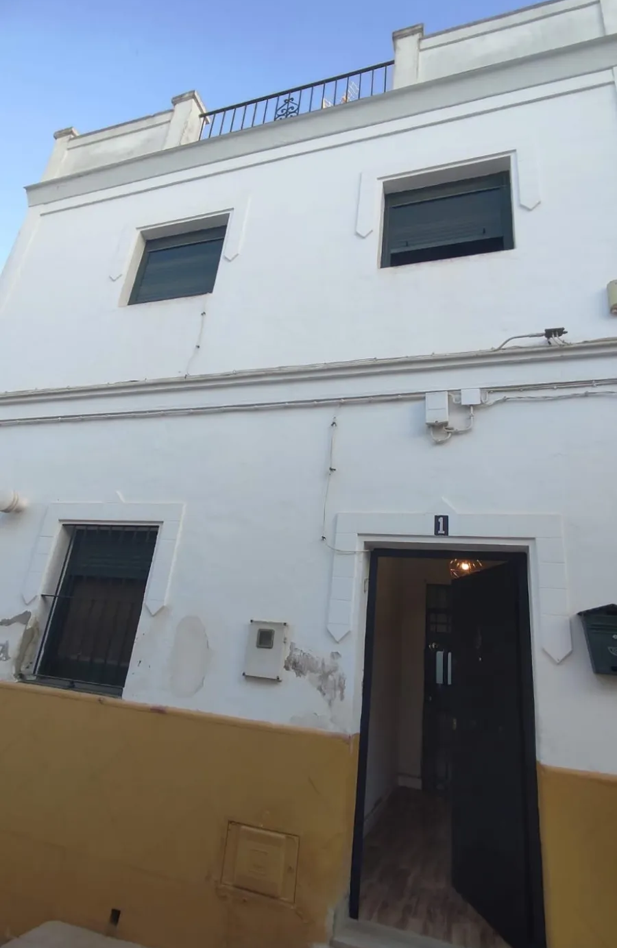 Edificio blanco de dos plantas con ventanas pequeñas y una puerta abierta, visto desde un patio estrecho.